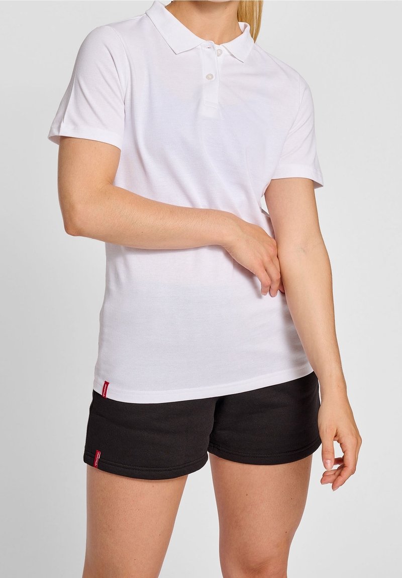 Personne portant un polo blanc à manches courtes et un short noir, ajustant la manche avec la main droite, debout devant un fond uni.