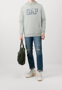 Sudadera con capucha gris con el logo "GAP", jeans azules desgastados, zapatillas blancas y marrones, y un bolso de piel sintética verde oscuro. Conjunto casual mostrado.