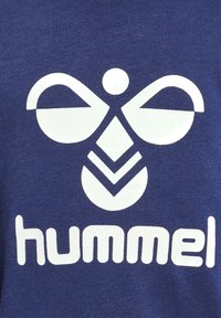 Niebieska koszulka sportowa z wyraźnym białym logo, zawierająca abstrakcyjne wzory oraz słowo "hummel" w grubym kroju pisma na dole.