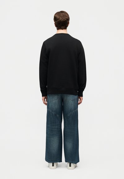 Άτομο που στέκεται με την πλάτη στραμμένη, φορώντας μαύρο μακρυμάνικο τοπ, φαρδιά μπλε τζιν και λευκά αθλητικά παπούτσια σε απλό λευκό φόντο.