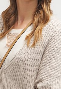 Cardigan en maille gris clair avec un col en V et une coupe ample, porté sur un haut blanc avec un détail en dentelle. Bandoulière marron visible.