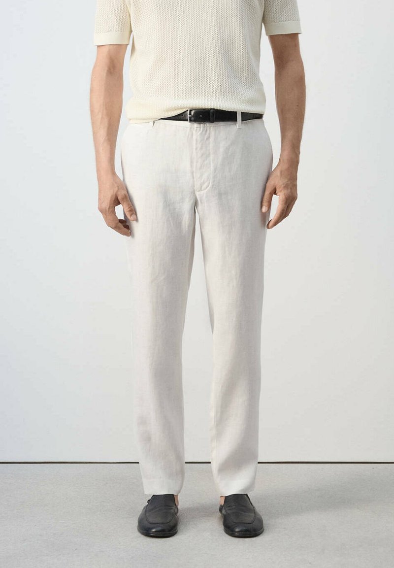 Homme portant une chemise blanche en maille à manches courtes, un pantalon blanc avec ceinture noire et des chaussures noires sans lacets, debout sur un sol gris.