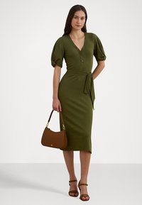 Olivgrön, knälång klänning med puffärmar, knappslå och bälte. Modellen håller en brun handväska och bär bruna klacksandaler.