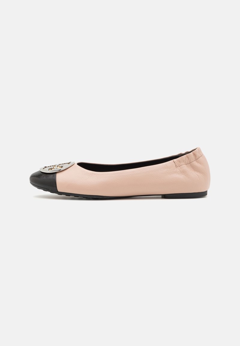 Ballerine rose pâle avec embout noir et emblème métallique rond à l'avant, talon élastiqué, et semelle plate en caoutchouc noir.