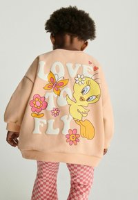 Fersken sweatshirt med hvid "LOVE TO FLY" tekst, prydet med en gul tegneserie fugl, lyserød sommerfugl og blomster. Teksturerede strækbare manchetter.