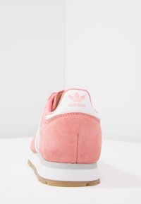 Rosa mockalädersneaker med vita accenter, med en texturerad ovandel, logotyp på hälen och en tan gummisula. Bakifrån med fokus.