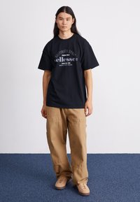 Svart grafisk t-skjorte med "ellesse"-logo, parret med løse beige bukser og lys brune sko på en blå teppebakgrunn.