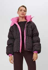 Žena v čiernej a ružovej prešívanej bunde so zvieracím vzorom leopardieho tlače, v kombinácii s tmavými nohavicami, stojaca na jednoduchom svetlom pozadí.