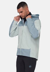 Homme ajustant la capuche d'une veste zippée gris clair et bleue, portant un pantalon noir avec un logo sur la jambe gauche, sur un fond uni.