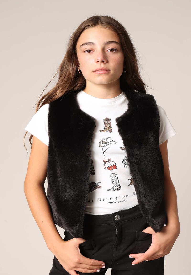 Gilet en fausse fourrure noire sans manches, texture douce, porté sur un t-shirt graphique blanc mettant en avant des bottes et des accessoires, associé à un pantalon noir.