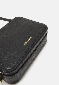 Sac en cuir synthétique noir avec motif crocodile, doté d'une fermeture éclair et d'un accent en logo doré. Forme rectangulaire avec une texture lisse et un léger éclat.