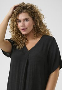 Haut noir ample avec un col en V et des manches larges, fabriqué en tissu texturé. Présente un pli avant pour un détail supplémentaire.