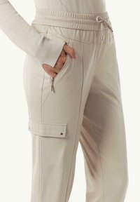 Beige joggingbroek met een elastische tailleband met trekkoord, een ritszak aan de zijkant en een cargozak met drukknoopsluiting. Glad, lichtgewicht materiaal.