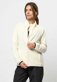 Vaaleanbeige fleece-takki, jossa on korkea kaulus, vetoketjukiinnitys sekä mustat reunat hihoissa ja helmassa. Takissa on huomaamaton logo rinnassa.