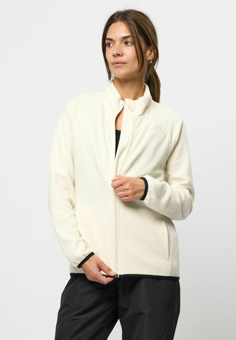 Vaaleanbeige fleece-takki, jossa on korkea kaulus, vetoketjukiinnitys sekä mustat reunat hihoissa ja helmassa. Takissa on huomaamaton logo rinnassa.