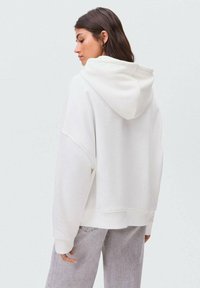 Felpa bianca oversize con un taglio rilassato e un ampio cappuccio, realizzata in tessuto morbido. Presenta polsini e orlo a coste, indossata con jeans grigio chiaro.