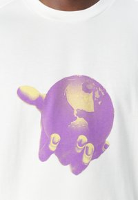 T-shirt blanc avec un graphique d'une main tenant un globe, comprenant des couleurs violettes et jaunes. Le globe montre des masses terrestres et des contours.