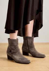 Botas de tobillo de ante gris con tacón cuadrado, que cuentan con una correa envolvente y una cremallera lateral. La textura parece suave y lisa.