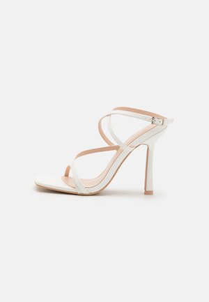 Biała sandałka na obcasie z cienkimi, skrzyżowanymi paskami i klamrą na jednolitym białym tle.