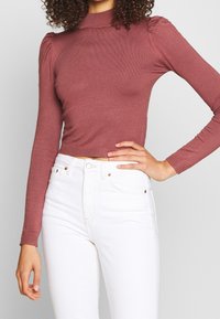 Haut à manches longues et ajusté en tissu côtelé doux de couleur bordeaux, avec col montant et détails froncés aux épaules. Associé à un jean taille haute blanc.