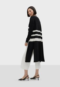 Svart lång kofta med vita horisontella ränder, tillverkad av mjukt tyg. Har en rundad halsringning och sidoflikar. Bärs med vita kortbyxor.