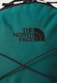 Mochila de tela en tono verde azulado con acentos negros, que presenta el logo de "The North Face" y un diseño de cordón elástico en forma de cruz en la parte frontal.