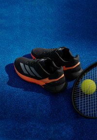 Μαύρα αθλητικά παπούτσια με πορτοκαλί λεπτομέρειες, με διαφανή υφή, τρεις ασημένιες ρίγες και ευέλικτη σόλα. Τοποθετημένα σε μπλε επιφάνεια.