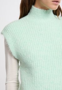 Maglione senza maniche di colore verde menta con collo alto, caratterizzato da una texture a coste e un materiale morbido e peloso. Lo strato interno è di colore chiaro.