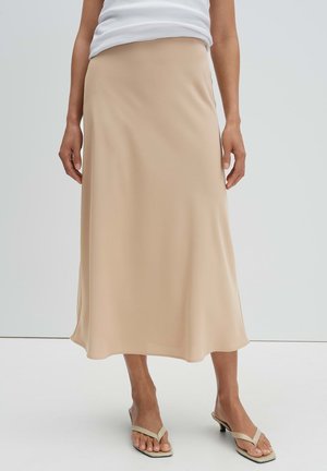 Vrouw draagt een beige midi-rok gecombineerd met beige sandalen met hakken en een witte mouwloze top, staand tegen een effen achtergrond.
