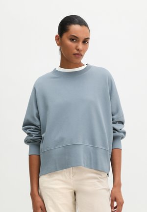 Vrouw met donker haar naar achteren gebonden, draagt een losse lichtblauwe trui over een wit shirt en lichtbeige broek, staand tegen een effen achtergrond.