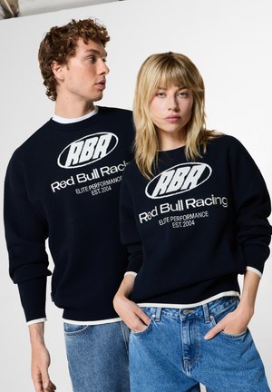 Zwei junge Erwachsene tragen passende schwarze Red Bull Racing Pullover und blaue Jeans und posieren vor einem einfachen weißen Hintergrund.
