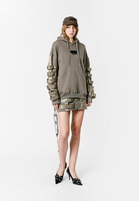 Sudadera caqui con paneles texturizados en las mangas, falda a juego y gorra negra. Presenta tacones de aguja negros brillantes y una etiqueta decorativa.