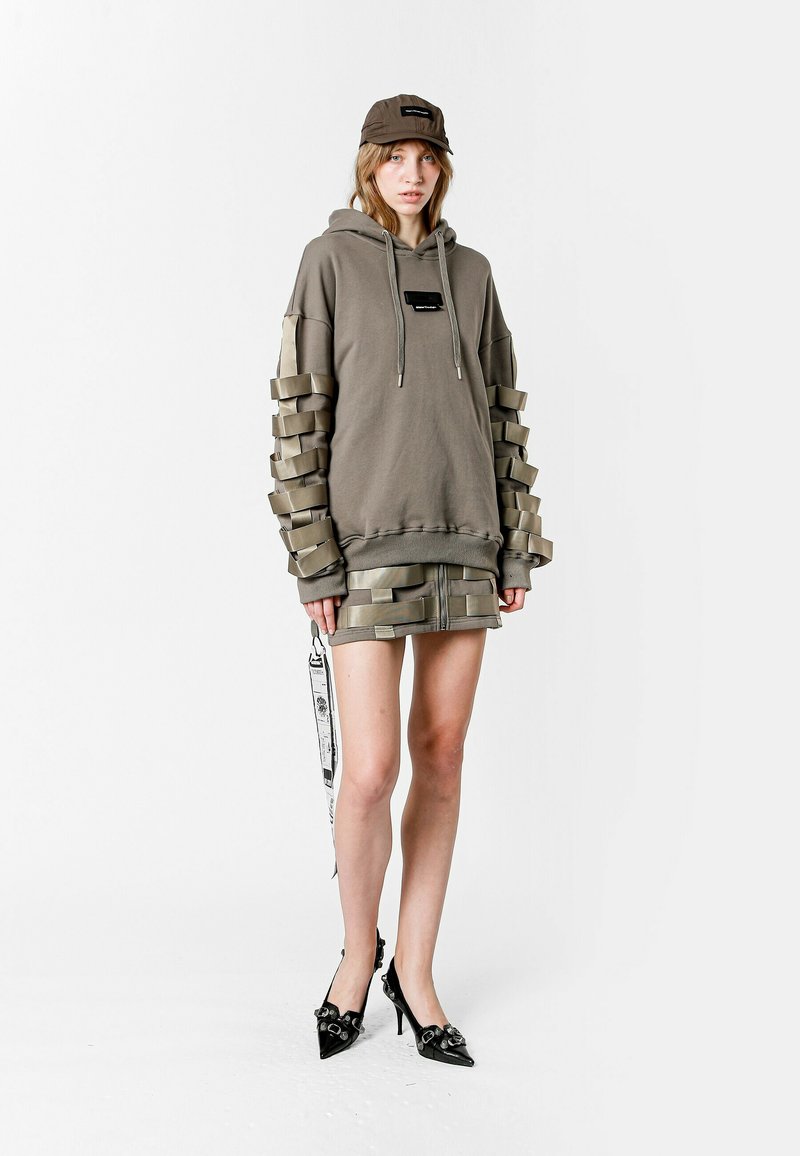 Sudadera caqui con paneles texturizados en las mangas, falda a juego y gorra negra. Presenta tacones de aguja negros brillantes y una etiqueta decorativa.