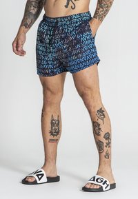 Plave i crne kupaće gaće s grafičkim uzorkom po cijeloj površini. Elastična waistband s uže. Nosene s crnim klompa sandalama koje imaju bijele akcentne.