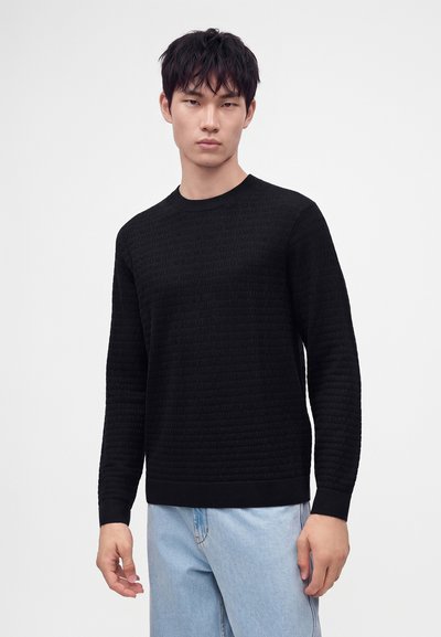Jeune homme avec un pull noir texturé et un jean bleu clair debout sur un fond blanc.