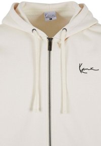 Cream hoodie with a zipper front, drawstrings, and an embroidered logo. Soft fabric with a smooth texture and black zipper accents.