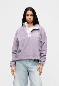 Μπλούζα από μοχέρ σε μωβ απόχρωση με γιακά μισού φερμουάρ, αντιθετικές κρεμ λεπτομέρειες και μπροστινή τσέπη με φερμουάρ· φορεμένη πάνω από ανοιχτόχρωμα τζιν με φθορές.