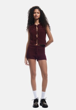 Ensemble en tricot bordeaux composé d'un haut court avec un nœud devant et d'un short à cordon. Porté avec des chaussettes blanches et des chaussures noires.
