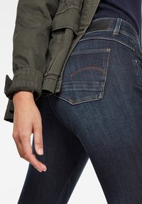 Jeans de mezclilla azul oscuro con un ajuste slim, que cuentan con bolsillos traseros delineados en costuras naranjas contrastantes. El material parece suave y elástico.