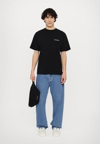 Svart bomullst-skjorte med teksten "No Problemo", kombinert med lyseblåt, løstsittende jeans. Modellen holder en liten svart veske. Hvite sneakers.