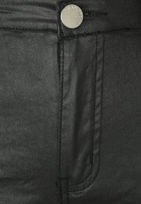 Tecido de ganga preto em close-up, apresentando um botão de metal com a gravação "GLAMOROUS", costura em contraste e detalhes do bolso frontal.