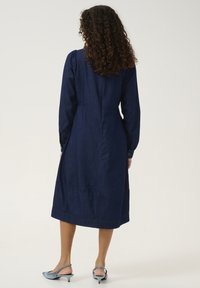 Marineblauw denim jurk met lange mouwen, een getailleerde taille, knoopmanchetten en een volle rok. De stof heeft een gladde textuur met minimale stikseldetails.