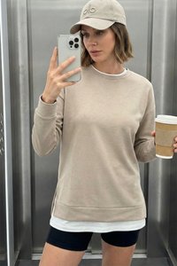 Kobieta w beżowej bluzie i czapce, czarne szorty, trzyma kubek z kawą i robi selfie smartfonem w windzie z metalowymi ścianami.