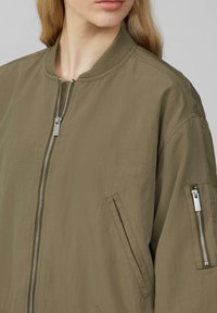 Olijfgroene bomberjack van lichtgewicht stof, met een rits aan de voorkant, zijzakken en een zak met rits op de arm. Soepele textuur.