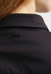 Czarna odzież z kołnierzykiem, o gładkiej fakturze. Z tyłu widoczna jest mała czarna metka z napisem "RAW". Prosty i minimalistyczny design.