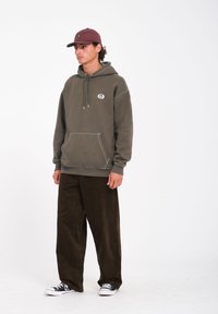 Olijfgroene hoodie met witte stiksel en een voorzak, gecombineerd met donkerbruine corduroybroek. Het model draagt een bordeauxkleurige pet en zwarte sneakers.