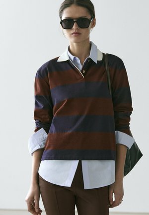 Femme portant de grandes lunettes de soleil noires, un chemisier à rayures bleu marine et marron à manches longues par-dessus une chemise à col bleu clair, et un pantalon marron.