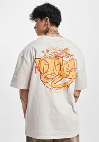 Cremefarvet bomulds t-shirt med korte ærmer. Stort grafisk print på ryggen med gule og røde abstrakte design, der inkluderer tekst og slangemotiver.