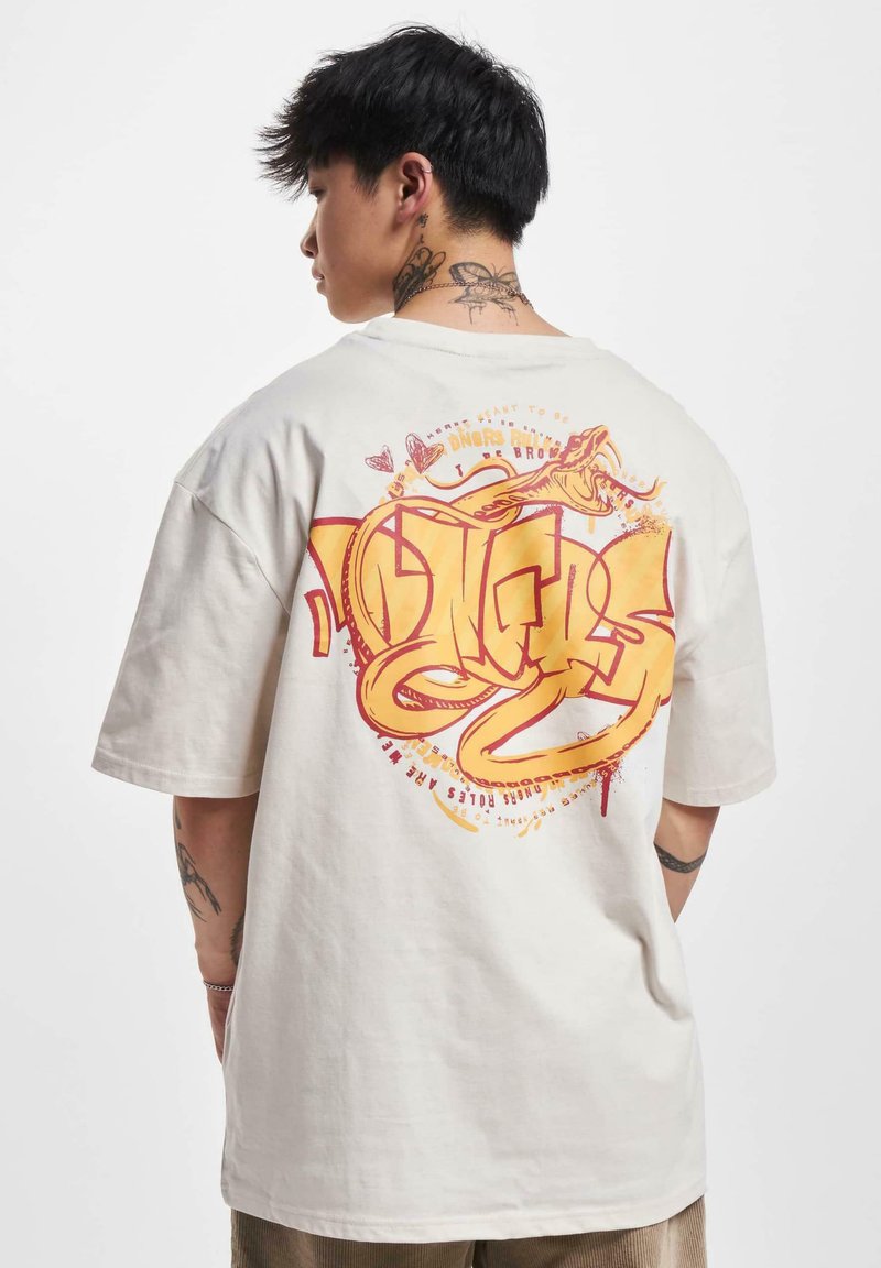 Cremefarvet bomulds t-shirt med korte ærmer. Stort grafisk print på ryggen med gule og røde abstrakte design, der inkluderer tekst og slangemotiver.