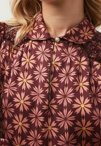 Ruskeissa kukkakuosisissa kankaissa on suuria vaaleanpunaisia ja keltaisia daisy-muotoja, joissa on tekstuurinen kaulus ja koristeellinen nappi.